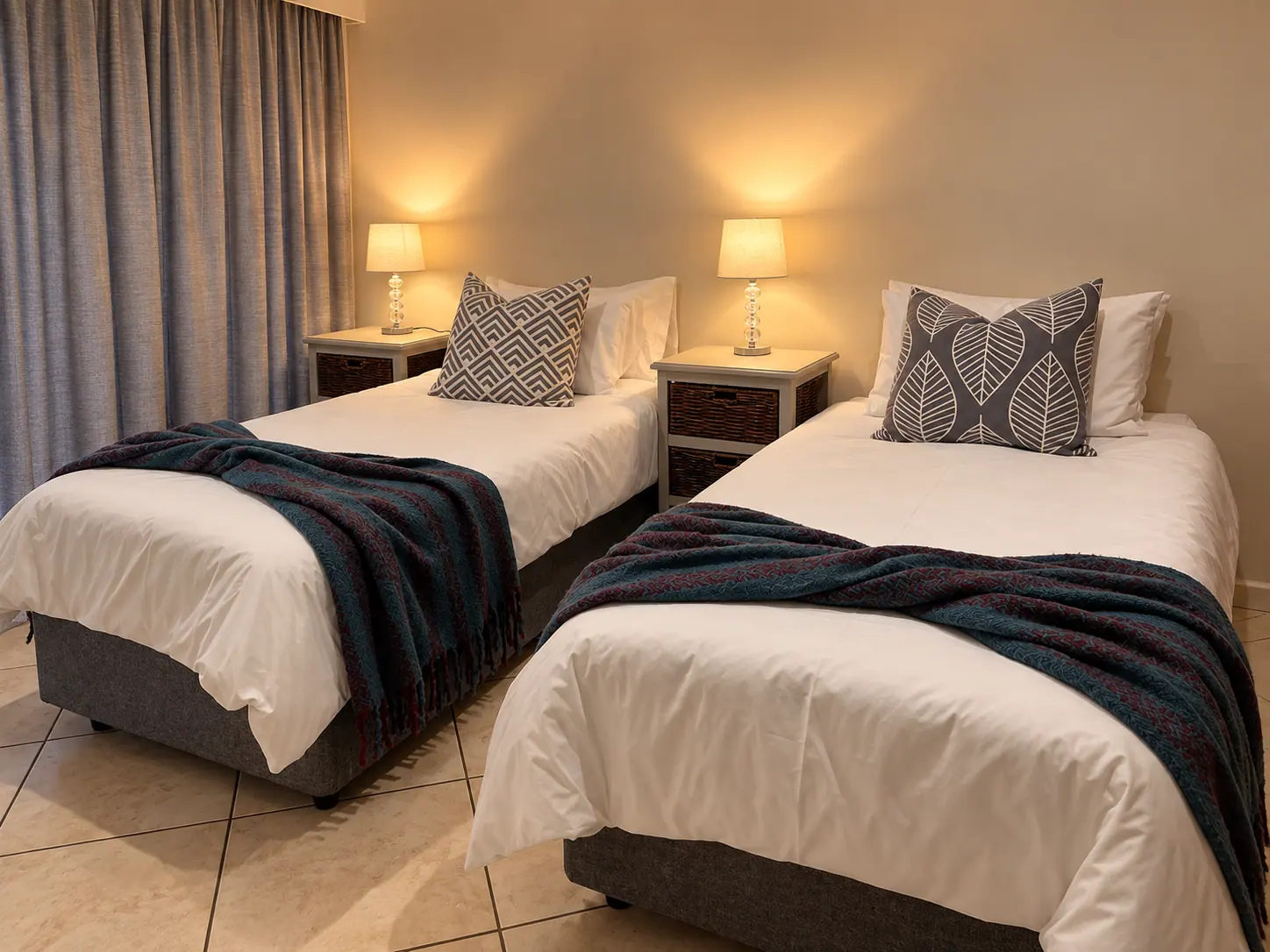 Second bedroom with twin beds, soft lamps, and blue-grey curtains